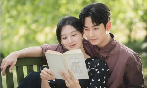 A couple is sitting on a bench, sharing a book in a green outdoor setting. The woman is smiling as she reads, while the man looks at her fondly. Both are dressed casually, with the woman in a polka dot top. From Kdrama "Queen of tears"