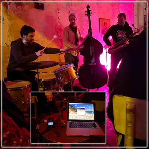 Ein Jazz-Trio in einem Jazzklub. Der Drummer (links) hat einen Drumstick im Mund und schlägt mit dem anderen auf die Cymbal. Rechts neben ihm steht ein Kontrabassist, der dem Schlagzeuger zusieht und seinen Kontrabass hält. Rechts davon steht ein Saxophonist und blickt nach unten. Die Band spielt vor einer rot angeleuchteten Wand. Hinter dem Drummer ist die Wand gelb. Am unteren Rand des Bildes befindet sich ein weißer Rahmen, in dem ein Laptop und ein Field-Recorder auf einem runden Tisch zu sehen sind.