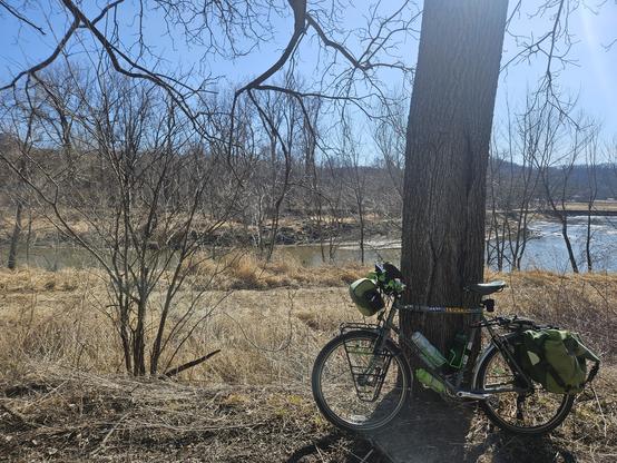 A bicycle with green bags leans against a tree. The grass is yellow and brown, a few small leafless trees are on the riverbank. A green river of water flows between the two riverbanks. The sky is blue.