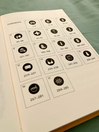 The book is opened and turned to the table of contents page. The contents page is designed as a grid of squares, each labeled with the chapter number and the page range of those chapters. For example, chapter 8 ranges from page 122 to 141. Also within each square is a black circle with a white icon inside each. The icons show different objects, such as a toaster, a bra, a bank, a bowl of cereal, etc.
