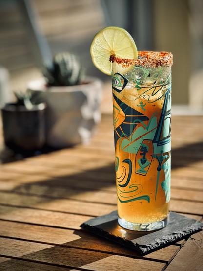 a light brown beer-based cocktail with red chili lime salted rim and fresh lime wheel sitting on a wooden table in the sun with potted cacti in the background