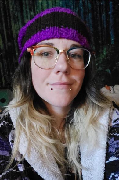 A person with long brown hair with blonde tips. They are wearing a purple and black hat, rainbow glasses with gold rims and have multicolored metal snakebite piercings. They are smiling. They are wearing a black, purple, and white top.