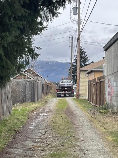 The tow truck is seen departing up an alleyway under a gray sky