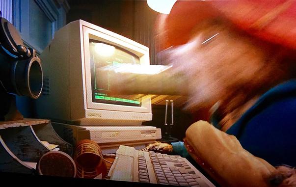 Shot from an unknown kids movie showing an Acorn computer of some sort on a desk with an animated beast of some variety with a large protuberance entering or exiting his mouth - the scene is confusing