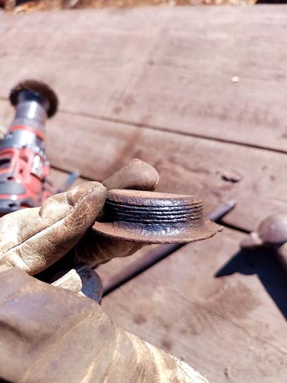 A threaded lid held in a gloved hand. The lid has a rusty thread that is somewhat perished and has been cleaned with a drill wire brush. The lid is also deformed, distorting the thread. It has two flanges for turning while the top remains flat.