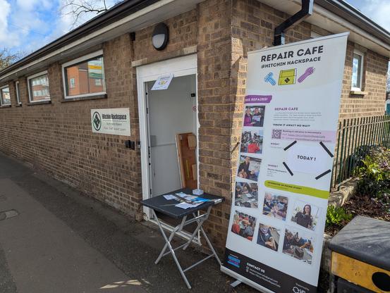 The open front door of the Hitchin Hackspace is in the centre of the image. The Hackspace is a single storey brick built building, a former public convenience. Just to the right of the door is a small square topped folding table with some handouts and flyers on it. To the right of the table is a "pop-up" banner about 2m tall with photos and information about the repair cafe events.