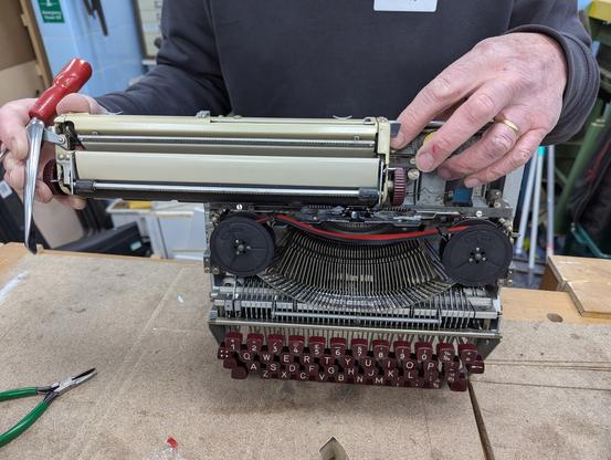 A mechanical typewriter in need of some tlc is stood on a workbench. It's outer casing has been removed and the person holding the machine is working on the carriage of the typewriter. Their fingers show signs that the typewriter may have had a nibble / resisted the efforts to fix it.