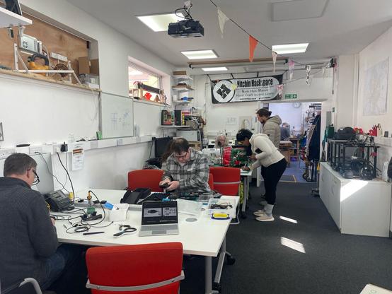 A general view of the inside of the Hackspace. On the left are pairs of tables next to the wall with repairers sat at the tables working on items. At a second pair of tables some visitors are watching a repair underway. Just behind them, on the right of the central gangway are the Hackspace 3d printers sat on their storage cupboards. In the background are the doors through in to the wood/metal workshop area of the Hackspace.
