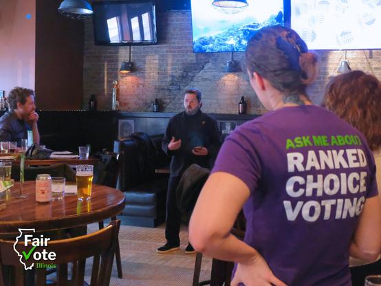 League of Women Voters of Chicago Vice President of Operations John Engle speaks about ranked choice voting at a bar in Chicago with supporters gathered around.