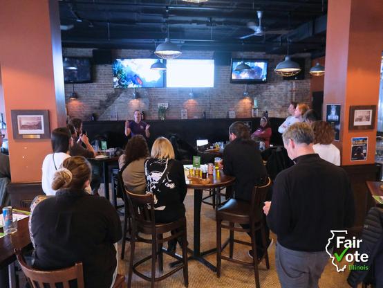 A large group of Chicagoans gather around for a presentation in a bar to learn about bringing ranked choice voting to their city.