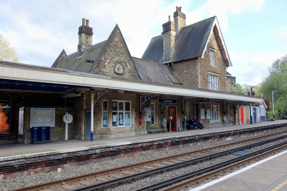 The image shows a daytime view of Godalming railway station in Surrey, England. The station building is constructed from light brown stone and features a steeply pitched, grey slate roof with multiple chimneys. The architectural style appears to be Victorian or Edwardian.
The platform is visible in the foreground, with two sets of railway tracks running parallel to it. The platform surface is a light grey, marked with a yellow tactile paving edge for safety. Overhead, a cream-coloured canopy supported by ornate metal pillars and brackets provides shelter.
Several doorways and windows are visible along the platform-facing side of the station building. Signage indicating "Godalming" is present above one of the entrances. A few people can be seen waiting on the platform.
Various station furniture and fixtures are present, including poster boards displaying advertisements and information, a roundel sign, and what appears to be a ticket machine.