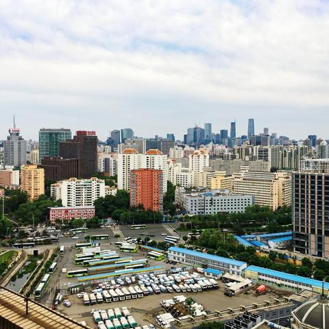 Aerial shot, a parking lot for trucks and a bus terminal in the foreground, residential highrises in the center, and the CBD in the background under a cloudy sky.