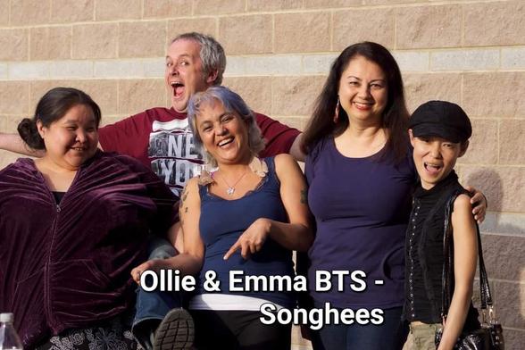 Medium shot of five people posing in front of a beige brick wall, evenly lit. A man, off-center to the left, is making a comical face. Four women surround him, all smiling broadly. Composition suggests a candid, informal setting. The text "Ollie & Emma BTS - Songhees" is superimposed on the bottom center of the image, providing context.