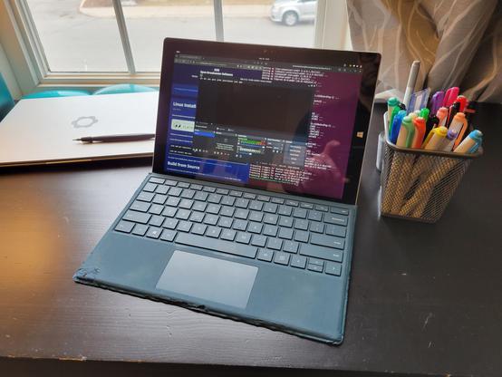 An old Microsoft Surface tablet, flanked by a closed Framework Laptop 13 and a cup full of pens and markers. Three windows are visible on the Surface's screen: the OBS website with installation instructions, an Ubuntu terminal window showing an OBS install, and OBS, running on top of them both.