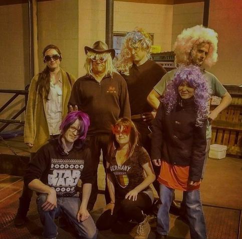 Medium shot of seven people in costumes, arranged in two rows. Lighting is somewhat dim and has a yellow cast. Bottom row: a person with a short purple wig kneels on the left, wearing a 'Star Wars' graphic hoodie with light denim jeans; a person with a red butterfly eye mask and a 'Team Germany' graphic t-shirt kneels next to them. Second row: Two women stand together and wear sunglasses, there is also a man who has on a cowboy hat and shoulder-length blonde wig and sunglasses. To the right, a man with a blonde wig, and lastly a woman with a purple wig, a black puffer jacket, a bright red skirt over blue jeans.