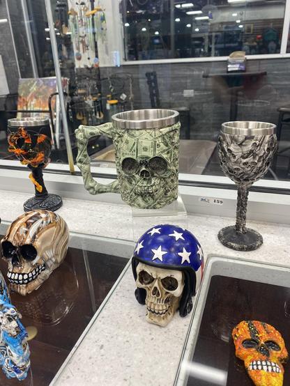 Several cups and statues of skulls sit on the glass surface of a cabinet.