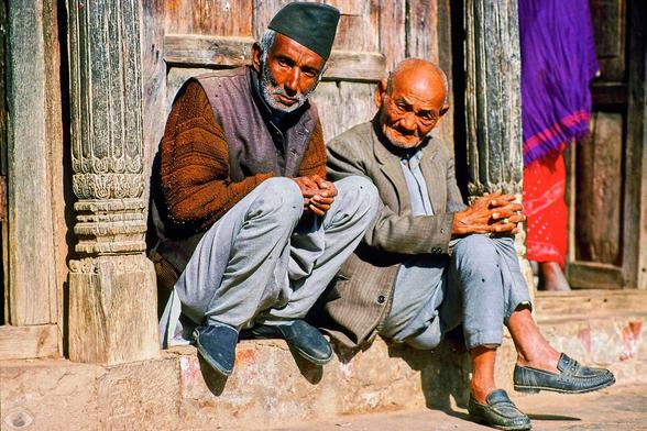 Two men sitting in front of a wooden facade of a shop in Nepal with a woman entering (only partly visible.) Both man are looking into the camera. One men in grey clothes is sitting on the threshold of the shop. The other men with grey pants and brown pull and black hat is crouched down on the same threshold.