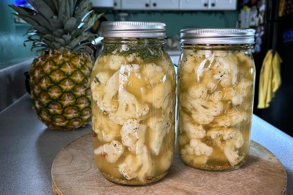 Two large glass jars sitting on a wooden cutting board filled with pieces of cauliflower in pickling brine and spices. There is a pineapple sitting on a counter in the kitchen behind the jars.
