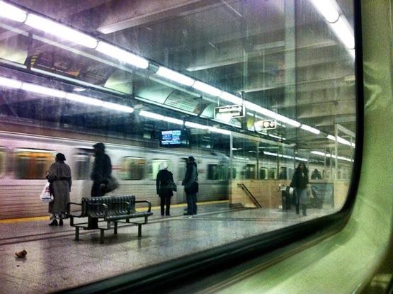 Looking out a window from a subway that has come to a stop.  We can see the platform between the tracks.  The subway from the other direction is slightly blurred, so either is just leaving or is slowing down.  Some people are standing facing it, indicating that perhaps it is just arriving.
