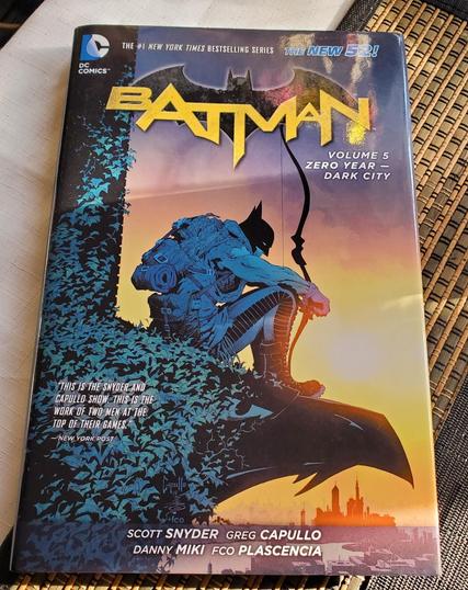 Comic book cover. Features Batman in silhouette, perched above the city skyline at sunset. The title "Batman" is in bold yellow, with "Volume 5, Zero Year - Dark City" beneath. The image is stylized with teal foliage and a gradient orange background. DC Comics logo in the upper left corner.