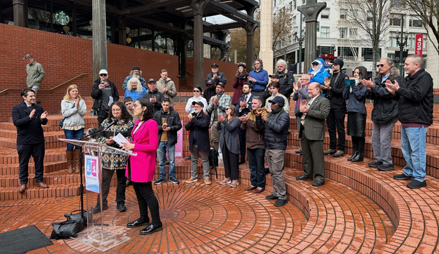 Two Genital Autonomy Collective Representatives giving a speech at a podium at the Pioneer Courthouse Square with a large group of Genital Autonomy Advocates clapping and standing behind on the round brick steps of the square.  
Me in a pink blazer, next to our science communicator in a geometric designed sweater in front of the Podium. The Podium has the Intact Global Icon consisting of pink and blue stylistic infants on a law scale at the same level indicating equality.