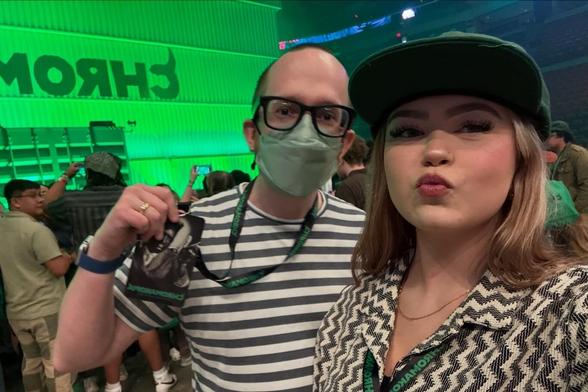 A man and woman take a selfie at an event with a bright green-lit stage in the background.