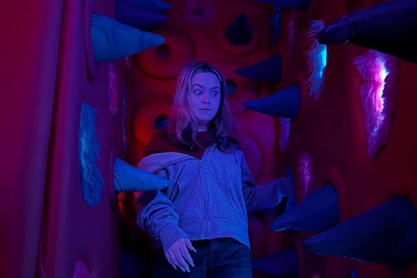 A woman stands cautiously in a narrow, padded tunnel with large blue spikes and glowing lights.