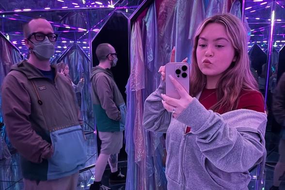 A woman takes a selfie in a mirrored room with purple lighting, while a man stands nearby wearing a mask.