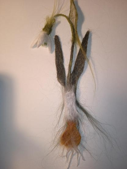 On a white background lies a snowdrop bookmark made from nettle using needle felting. It is not yet finished, the parts of the bookmark are superimposed on each other so that you can understand how it will look when it is finished. Thank you for your attention!