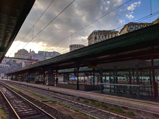 A ground-level, eye-level shot shows the multiple platforms and tracks of Genova Piazza Principe train station in Genoa, Italy, under a cloudy sky with hints of sunlight. The station has multiple parallel platforms covered by dark green, ornate metal canopies supported by dark metal pillars. Each platform has a light-coloured surface with a yellow safety line near the edge. Some platforms have enclosed waiting areas with glass walls and metal frames. Several sets of parallel train tracks run along the ground between the platforms, showing gravel and some sparse vegetation between the sleepers. Overhead power lines stretch across the scene. In the background, numerous multi-story buildings in varying shades of beige and brown are visible, built on a slight incline. The sky is predominantly grey with patches of blue and soft yellow light peeking through the clouds near the horizon.