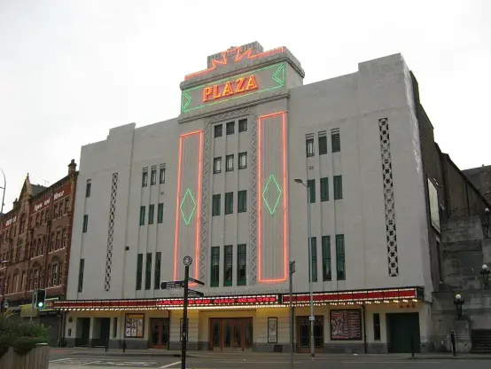 The image features the exterior of a building, specifically a theater or cinema. It has a distinctive Art Deco design, characterized by its vertical lines and decorative elements. The marquee displays the name "PLAZA" prominently, and the facade is adorned with neon lighting and geometric patterns. The overall appearance suggests it may be a historic venue that has been preserved or renovated.

Image Credits: Wikimedia / Stephen Armstrong  / CC BY-SA 2.0