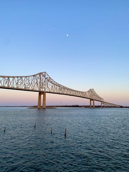 A long cantilever bridge stretches across a wide river under a sunset sky. Near the horizon, the sky glows light purple, gradually shifting to hues of orange, yellow, and blue as it rises. The bridge has a soft orange glow, reflecting the light of the setting sun.