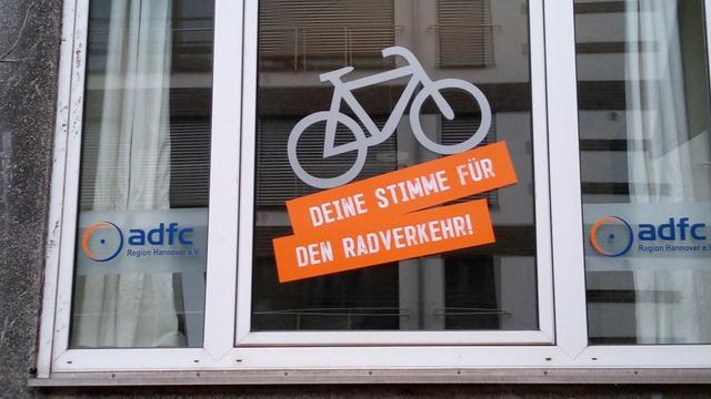 Fensterfront des ADFC-Infoladen mit neuer Beschriftung. Ein aufwärts fahrendes Fahrrad in weiß mit Schriftzug drunter "Deine Stimme für den Radverkehr" in weiß auf orangem Grund. Links und rechts in kleinen Fensterflügeln das Logo des ADFC mit dem Schriftzug "ADFC Region Hannover"