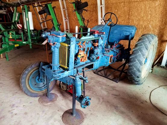 An old blue tractor that has been heavily modified to serve as teaching aid. Parts are painted in highlight colours, one wheel is removed, it rests on axle stands and a lot of holes have been cut into things to expose their inner workings.