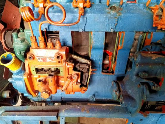Close-up of the four cylinder diesel engine. The cylinders are cut open, revealing the piston heads inside. One piston head is cut open too, the cut painted in red, revealing the pin and connecting rod. The fuel pump and two injector pipes are attached to the side, cut open in two places as well, painted in orange.