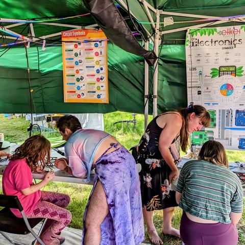 Two people dressed in femme outfits leaning in opposite directions each helping two younger kids with a soldering project on folding tables outdoors in a pop-up tent. Behind them hang colorful posters talking about electronics.