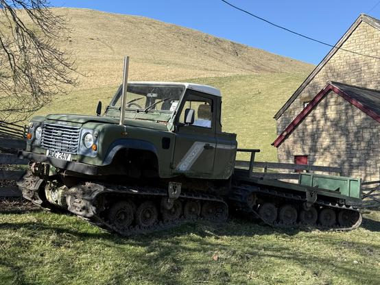 An off-road vehicle with tank tracks, painted green, is positioned near a stone building in a grassy landscape. The vehicle features a prominent exhaust pipe and a flatbed, set against a backdrop of rolling hills.