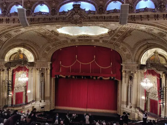 The image depicts the interior of a theater. It features an ornate design with elaborate moldings, chandeliers, and a grand stage draped in red curtains. The architecture appears to be classical, with intricate details on the ceiling and walls. The view shows some audience seating and a few people gathered, likely before a performance.

Image Credits: Wikimedia / Sdkb / CC BY-SA 4.0