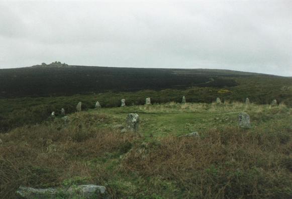 A large part of the circle of standing stones of the eastern, and most complete, of the two stone circles on the edge of the moorland common by Tregeseal in St Just in Penwith parish in West Cornwall. The stones stand in a vague clearing of brighter green grass surrounded by the rougher moorland plants. Some of the ground in the vicinity has been burned. The impressive tor of Carn Kenidjack rises to the horizon on the left. The sky is steely grey.