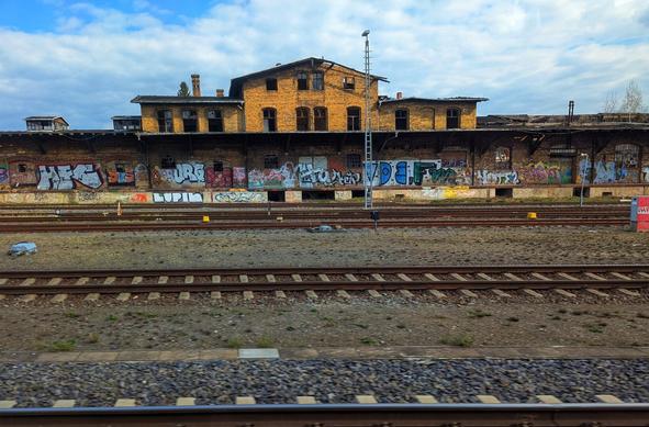 The image shows a slightly elevated view across multiple sets of railway tracks towards a derelict, two-storey brick building under a partly cloudy sky. The building is constructed from yellow-brown bricks and features numerous arched windows, many of which are boarded up or broken. The roofline is uneven, suggesting disrepair, with some chimney stacks visible.
Attached to the front of the brick building is a long, lower structure, possibly a former platform, which is heavily covered in colourful graffiti. The graffiti spans the length of this section, featuring various styles and lettering. Below this platform area is a patch of rough ground with some sparse vegetation.
Several sets of parallel railway tracks run across the foreground and middle ground of the image, suggesting a former or current railway station. The tracks are laid on gravel and sleepers.