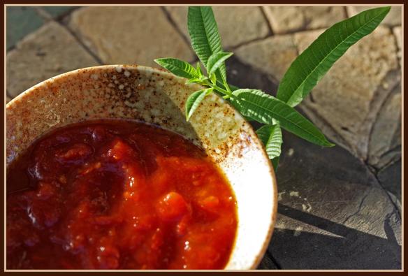 A tomato sauce sits in a mottled pottery bowl on a stone table with a sprig of lemon verbena.