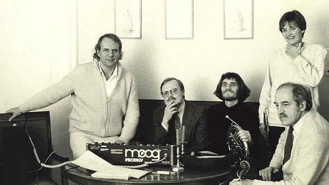 A black&white photo of five people gathered around Moog prodigy synthesizer on a table with a few papers, cables, and equipment.
People are: Karlheinz Stockhausen (standlng/leaning), Jørgen Lekfeldt, Carl Bergstrøm-Nielsen, Lene Maack (standing), and Ivan Vincze.