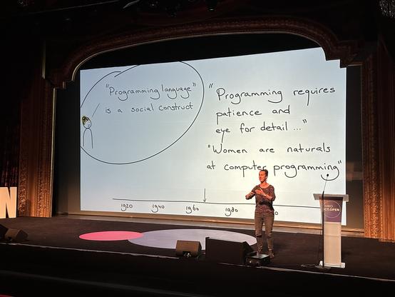 Felienne presents a slide in which a cartoon version of herself says "'Programming language' is a social construct", and presents a timeline where the 1960s are labelled with two quotes: "Programming requires patience and eye for detail ..." and "Women are naturals at computer programming".