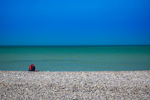 Horizontal dreigeteiltes Bild mit Himmel, Meer und Strand. Eine entfernte sitzende Person ist von hinten abgebildet.