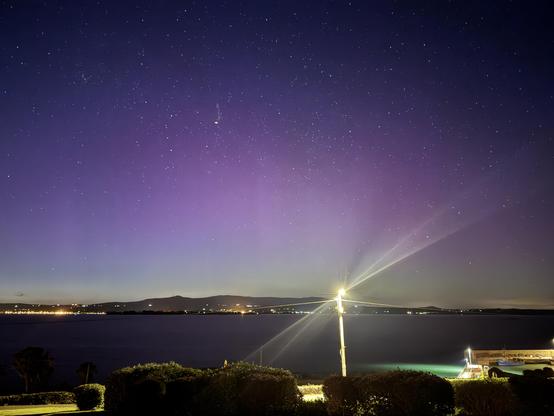 Aurora over Dungarvan bay looking towards the Comeragh Plateau from Helvick. An LED streetlight is flaring in the foreground. 

Lots of purple and green covering most of the sky.