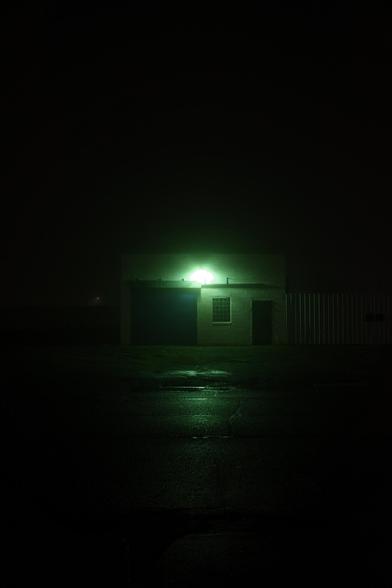 Color photograph looking across a darkened street on a foggy night at an old auto shop-like building. The face of the building with a closed garage bay door on the left, a block glass window in the middle and a darkened entrance on the right, is visible as if barely emerging from the darkness behind it, catching a greenish illumination from a very bright security light shining into the fog, reflected along the wet street in the foreground