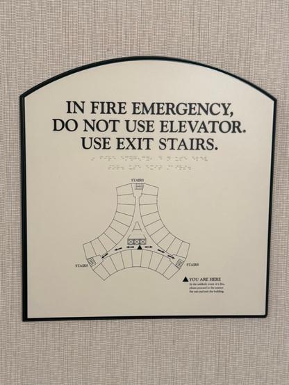 Fire escape floor plan that reads "IN FIRE EMERGENCY, DO NOT USE ELEVATOR. USE EXIT STAIRS." With braille below. It shows the curved triangular layout of the hotel with three intersecting hallways around the central core and rooms on the perimeter