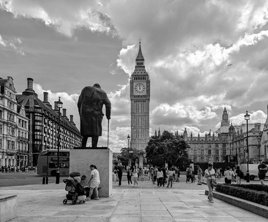 A black and white photograph captures a bustling scene in London, with the iconic Elizabeth Tower, commonly known as Big Ben, standing prominently in the mid-ground. The tower's clock face is clearly visible, and its intricate architectural details are highlighted by the monochrome tones.
In the foreground, a statue of Winston Churchill, depicted from the rear, stands atop a plinth. He is shown wearing a coat and holding a cane. A woman in a light-coloured top and shorts stands near the base of the statue, seemingly looking at it. A pram is parked beside her.
Numerous people stroll across the paved area in front of the statue and towards the Houses of Parliament, which are visible to the right of Big Ben. A red double-decker bus is partially visible on the left side of the frame, moving along a street lined with tall buildings featuring ornate architectural details.
The sky above is filled with dramatic clouds. A small aeroplane is visible in the distance.