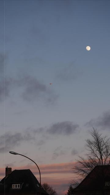 The image shows a landscape at dusk or dawn with a few houses, visible rooftops, and a streetlight. Above, the sky is partially cloudy with the moon visible. There's also a small helicopter flying in the sky. The lighting suggests a serene and peaceful view of a suburban or residential area.