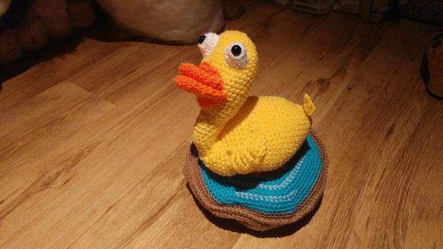 A yellow crochet duck with a big orange beak and large, googly eyes, sitting on top of a crochet puddle with a blue surface to indicate water and brown underside to indicate earth.